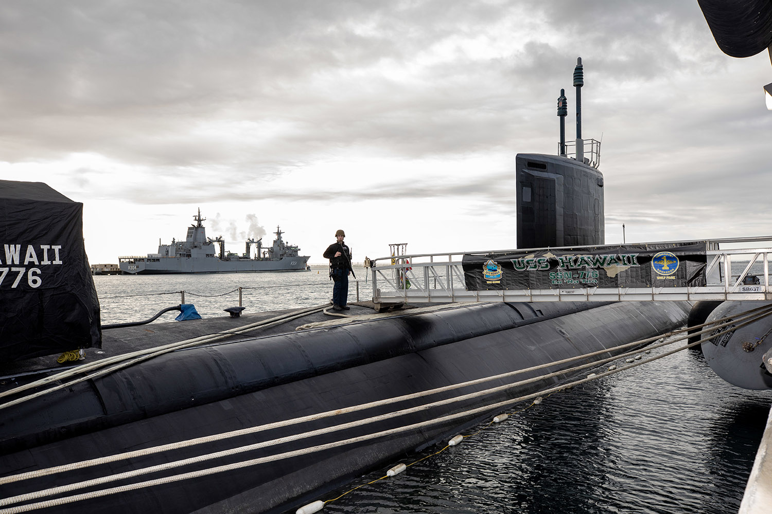 A sailor is seen from a distance, standing atop a submarine as it floats in the water. A large battleship can be seen in the background against a gray cloudy sky.