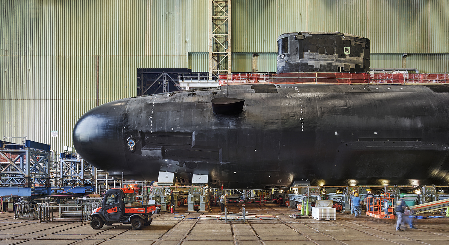 A rounded section of the hull of a giant submarine is seen under construction in a large building. Scaffolding hangs from parts of the black surface of the vessel while workers in hard hats bustle around on the floor below it.