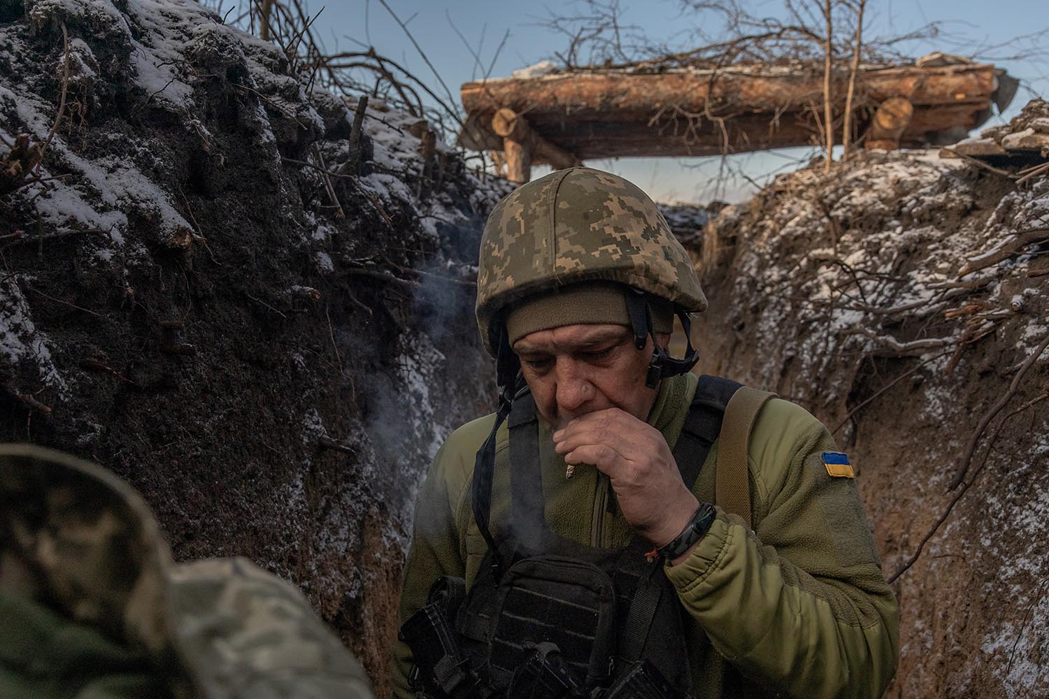 A soldier in a helmet and combat gear smokes a cigarette in a trench.