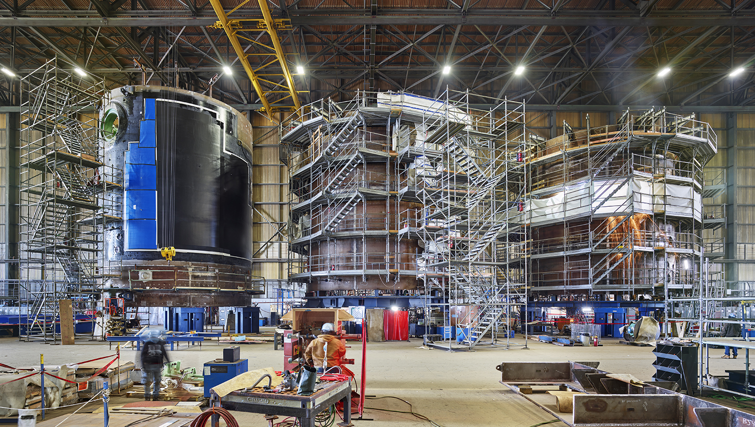 Three large towers of scaffolding surround cylindrical sections of a submarine under construction in a massive open room of an industrial building.