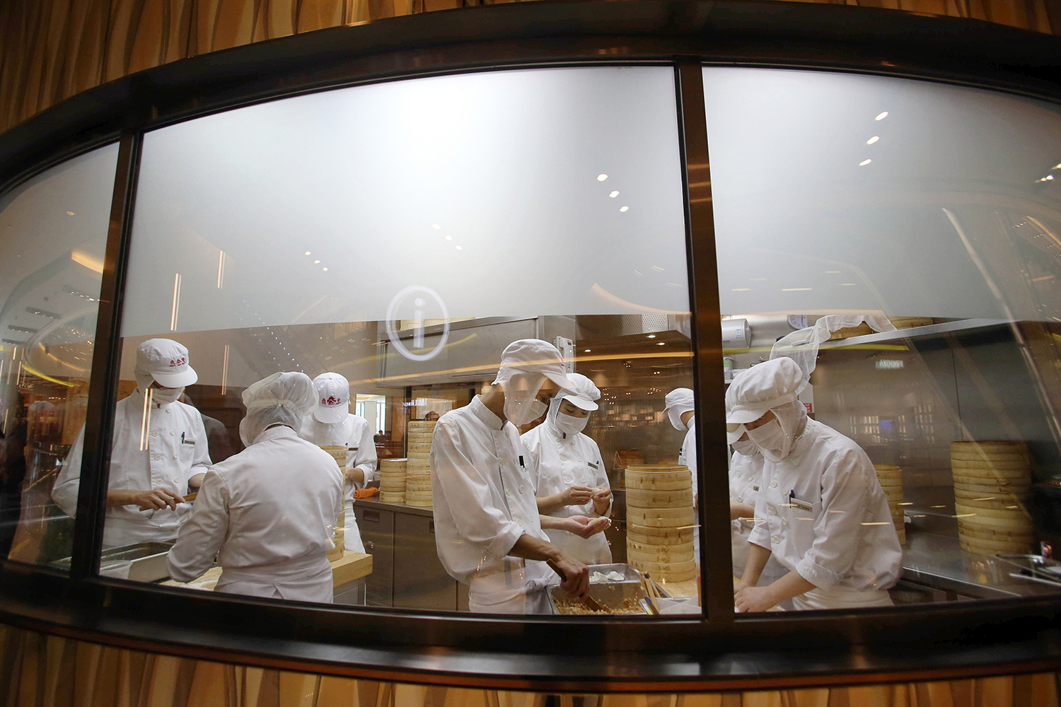 Workers in white smocks, hats, and masks are seen through a window making dumplings in a restaurant kitchen.