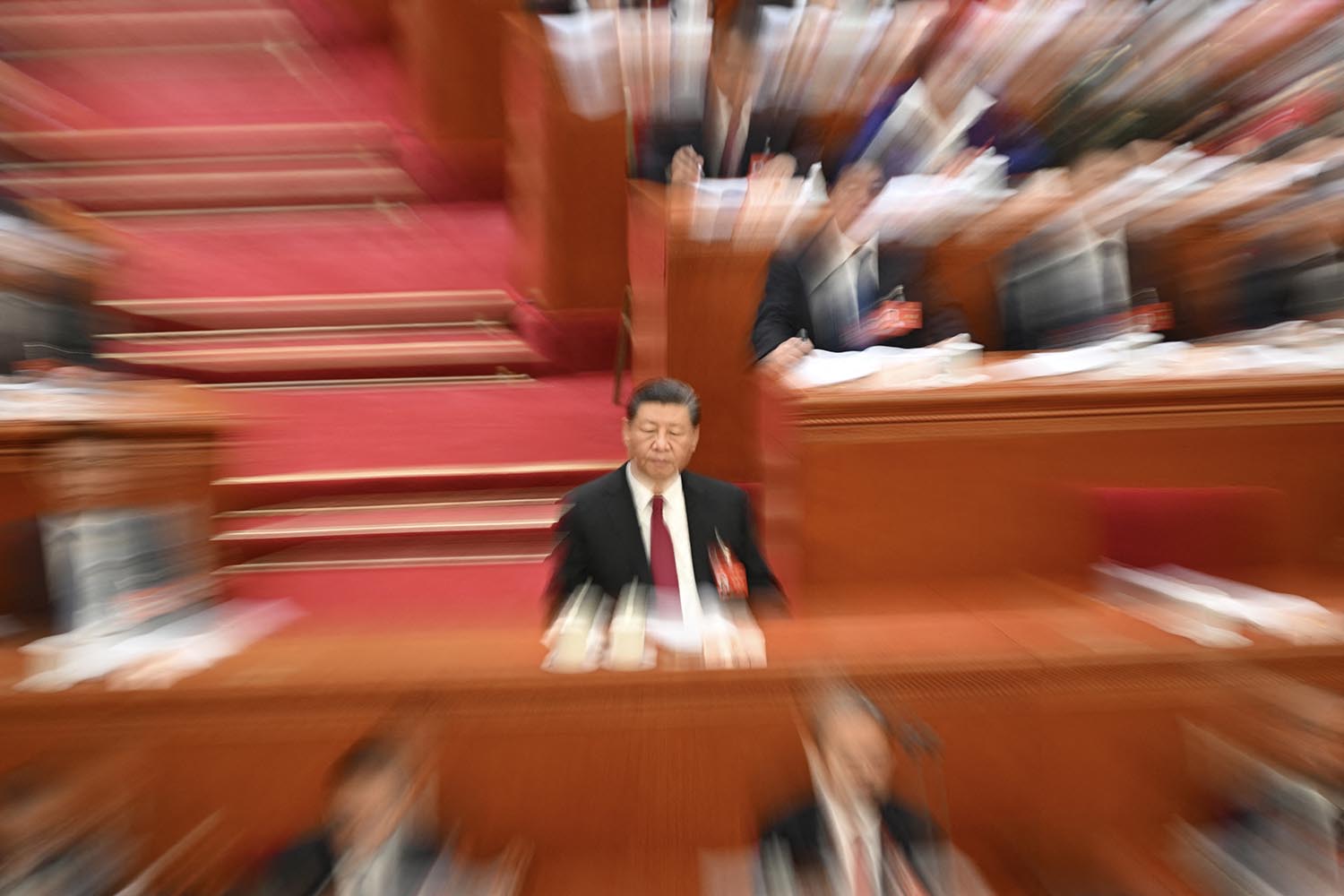 Xi Jinping sits at a long table. Other congressional members sit around him in blurry focus.