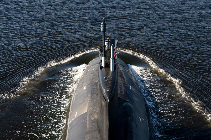 A U.S. flag and two people can be seen perched atop a portion of a partially-submerged submarine cutting through open water at sea, sending waves in motion behind it as it moves.