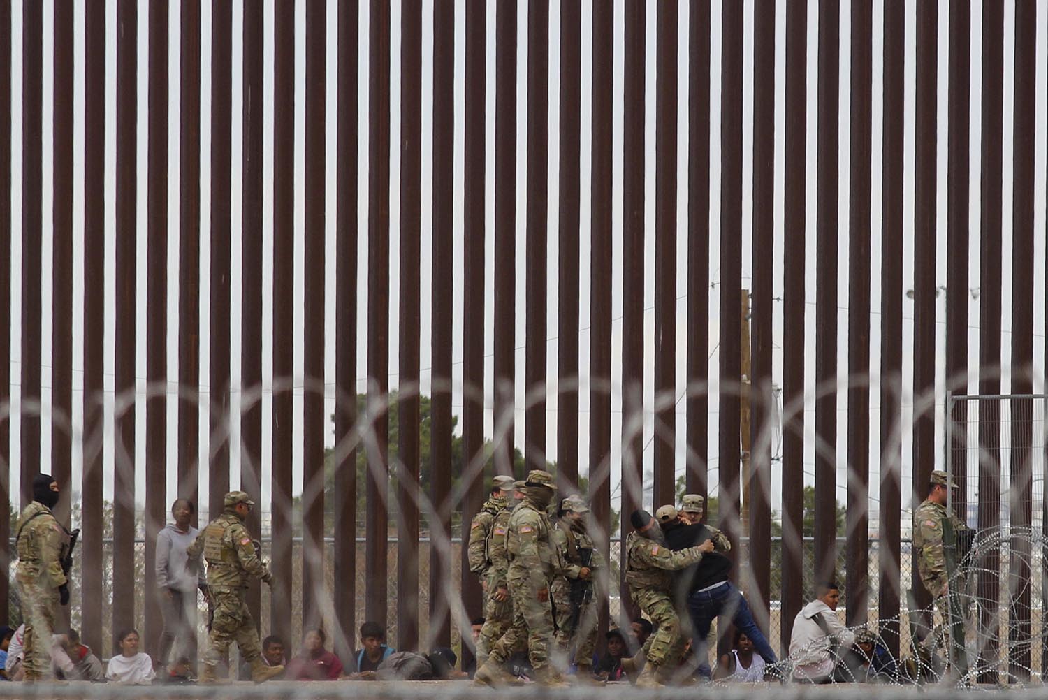 The tall slats of a border fence are in the background as several guards detain an immigrant, seen through razor wire.