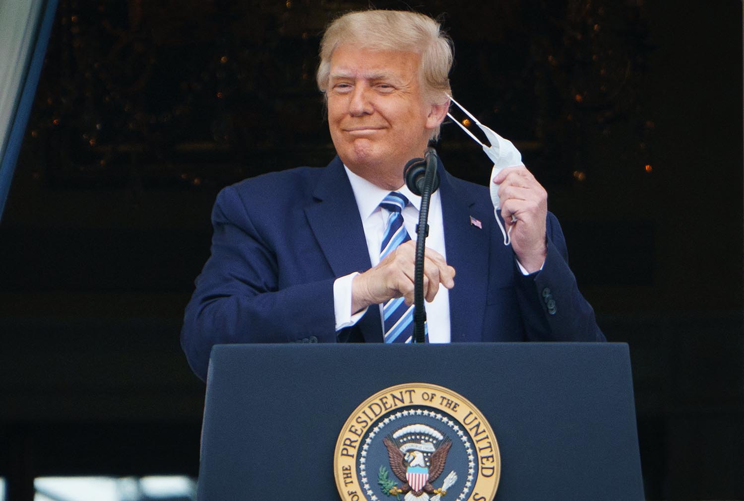 Donald Trump stands at a podium while taking off a face mask.