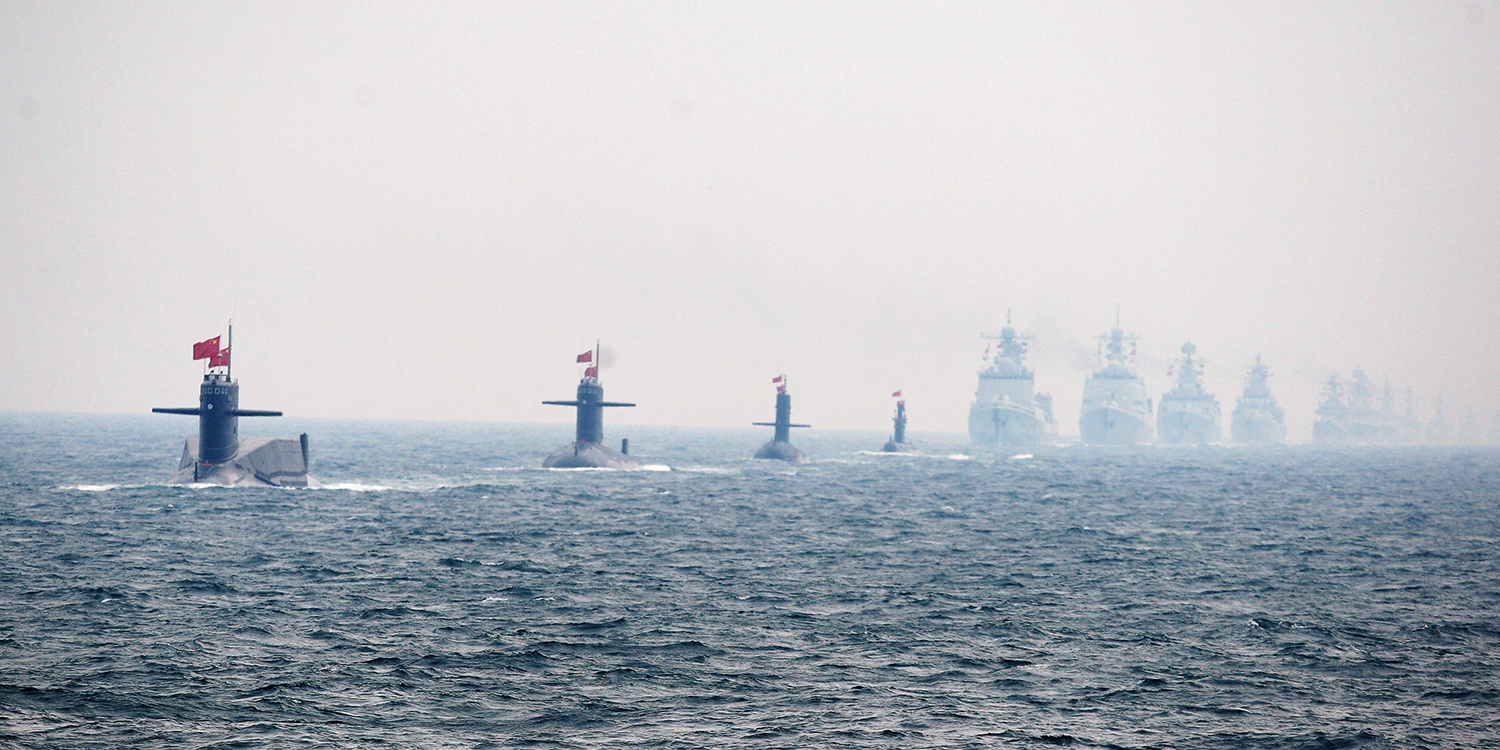 A neat line of four Chinese warships, with four submarines also lined up in the foreground, are seen in the open ocean. The more distant warships are partly obscured by fog beneath a hazy gray sky.