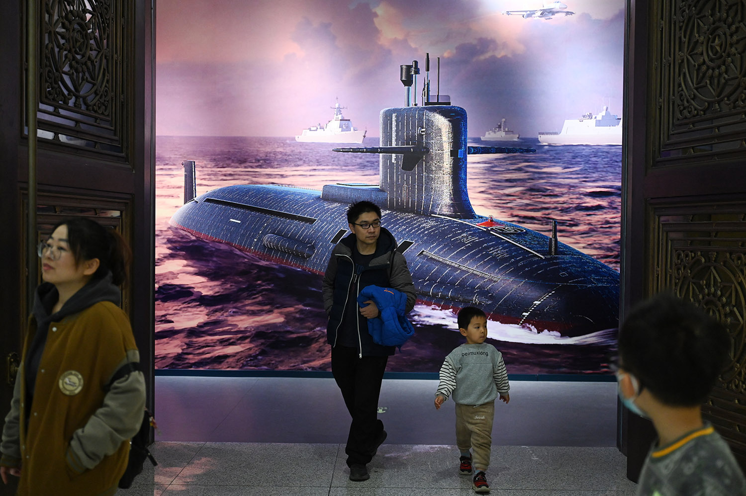 A young child and a man holding the child's jacket walk in front of an illustration of a submarine on the that hangs on a wall behind them. A few other people are seen in the room is they examine other exhibits.