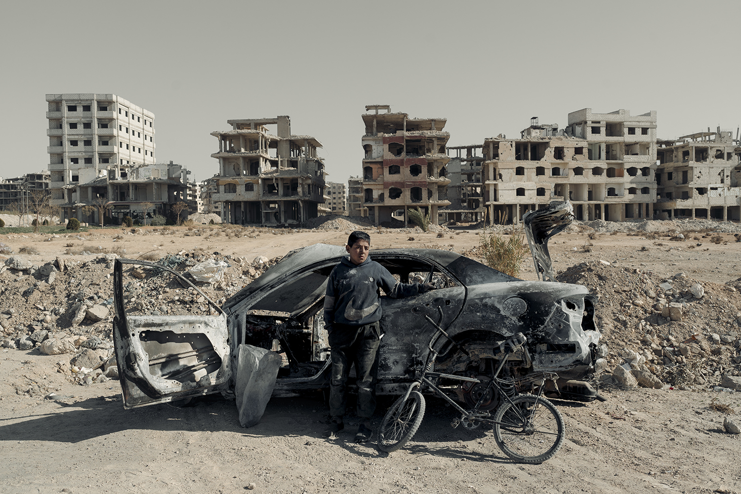 A boy stands in front of the shell of a bombed-out car on a packed-dirt street. A battered bicycle leans against to the car. Behind him are the war-torn shells of multistory buildings.