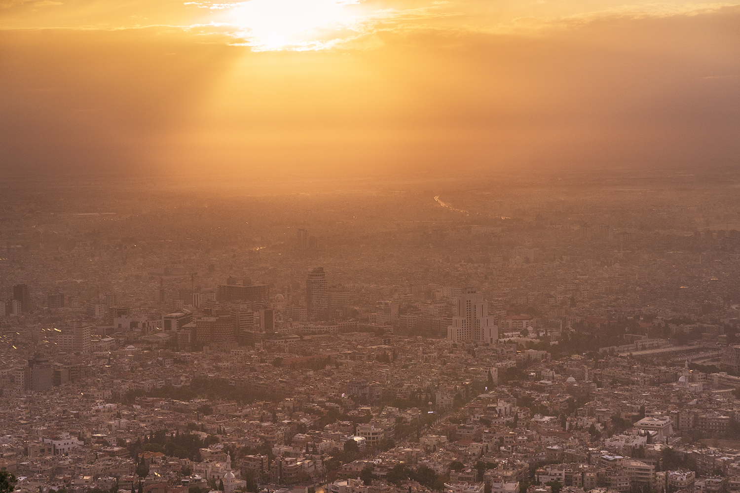 An elevated view shows the sunrise casting a glow on the white buildings of a broad cityscape below that extends to the horizon.