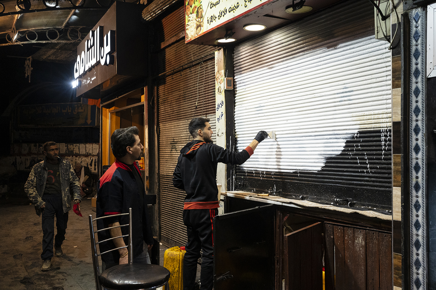 A man uses a paintbrush to use white paint to cover a metal sheet covering the front window of a business. Two other men, one walking by smoking a cigarette, and one leaning on a chair, watch on a darkened street with illuminated signs over businesses.