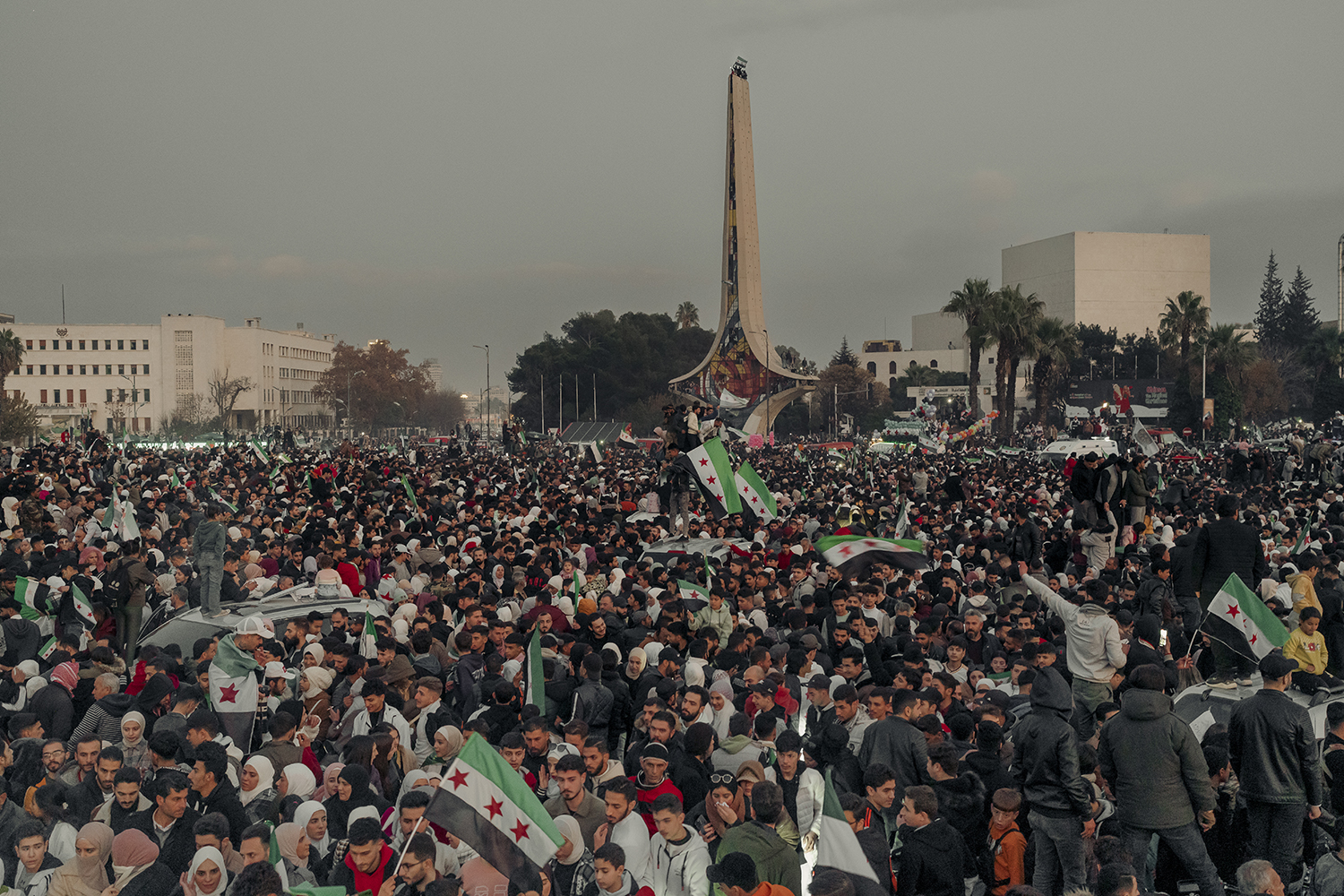 A crowd of hundreds of people fills a public square with a large monolith looming against a hazy sky in the background. Many people hold flags with a green, white, and black stripe with three red stars, a symbol of Syrian independence.