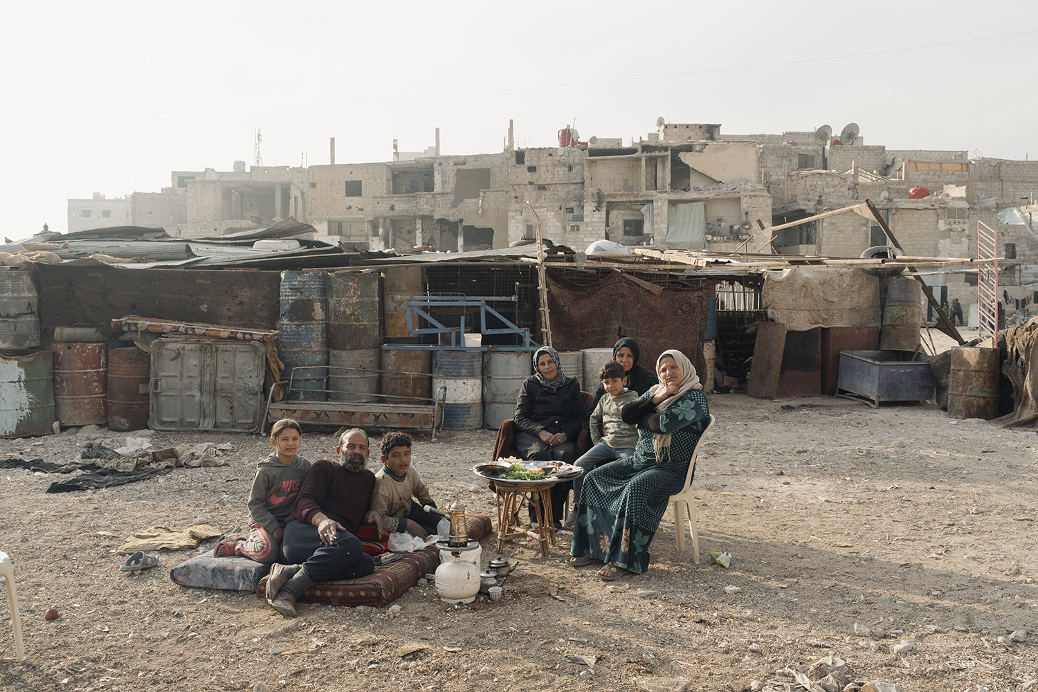 A man and two children sit on a mattress on the ground beneath a hazy sky next to a pot over being warmed by a propane tank. Three women and a child sit at a small table next to them with a platter of food on it. They are outside what appears to be makeshift shelters in front of war-damaged buildings.