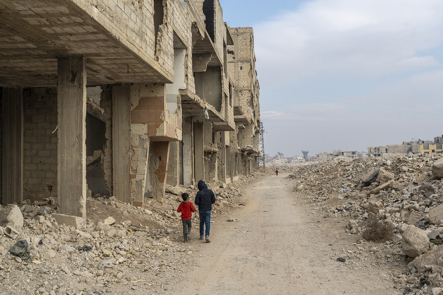 Two children walk along the edge of a dirt road. At their left is the shell of a building. At right, piles of rubble beneath a blue sky mostly covered with clouds.