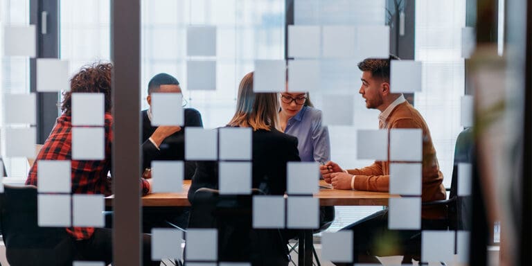 View through a glass panel with sticky notes, of a group of employees meeting in a conference room