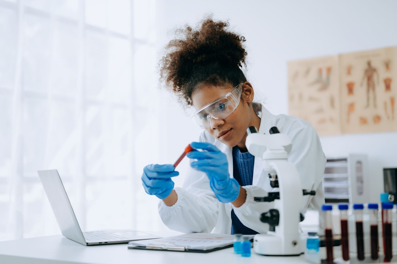 A woman dressed in a white lab coat examines a vial while sitting in front of a computer.