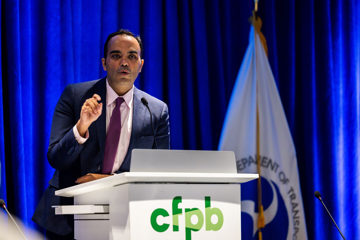 CFPB director Rohit Chopra speaking behind a podium with the green CFPB logo.