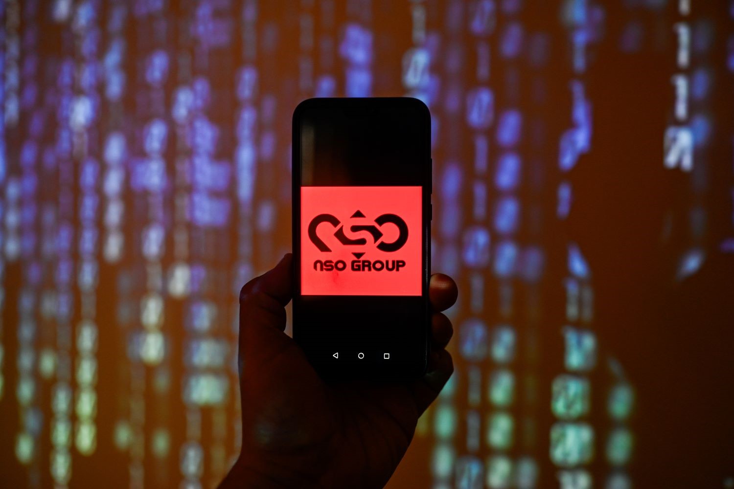 A shadowy hand holding a phone displaying the NSO Group logo.