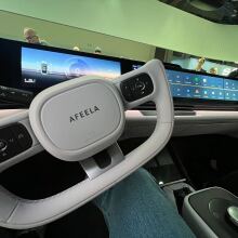 A close-up view of the interior of an AFEELA car, focusing on the futuristic steering wheel with "AFEELA" branding. The dashboard features a wide digital display with driving information and an app interface.