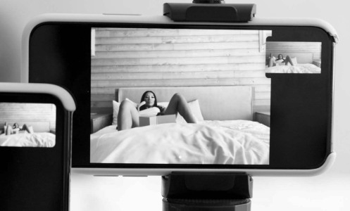 An iPhone sits on a tripod, filming a woman who lays with her legs open on a bed.