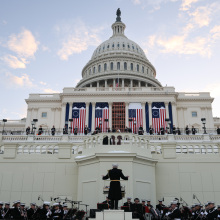 The United States Marine Band rehearsing their performance in front of the Capitol building, as large American flags drape across the facade.