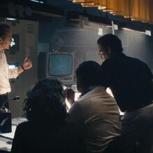 People sit and stand in a newsroom, talking and looking at one another. 