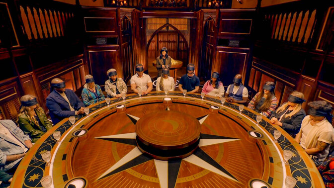 A group of people sit blindfolded at a large round table.