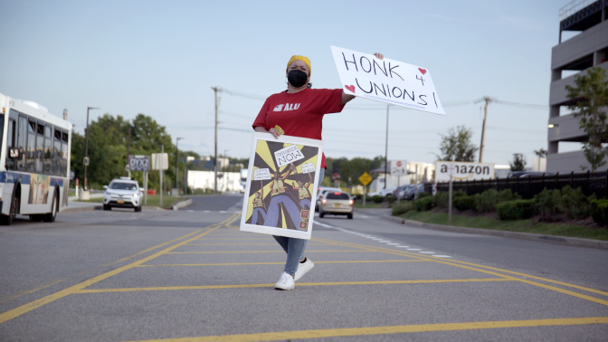 An ALU member stands in the median of a road holding a sign that reads "Honk 4 Unions."