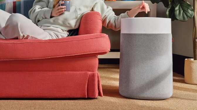 a blue air air purifier sits beside a woman sitting on a red couch