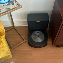 iRobot Roomba Combo j5+ and self-empty dock sitting against wall with yellow chair and TV stand in peripherals