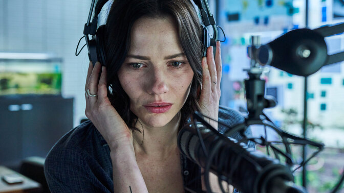 A woman sits at a desk in front of a microphone, wearing a headset.