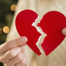 A person holds a paper heart with a rip down the middle, with a Christmas tree in the background. 