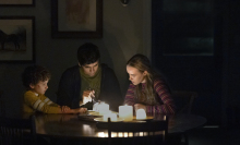 A family sit in their home in the dark, at the dining table lit by candles.