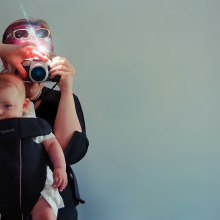 Still from 'Motherboard' shows Victoria Mapplebeck holding her baby son Jim in a sling carrier while she holds a camera to photograph them.