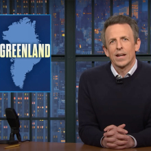 A man sits behind a talk show desk. In the top-left is an image of another man and a map of Greenland.