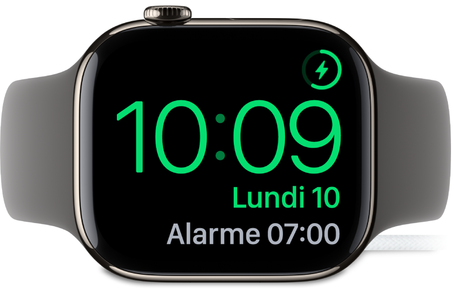 Apple Watch placée sur sa tranche et connectée au chargeur. L’écran montre le symbole de recharge dans le coin supérieur droit, l’heure actuelle en dessous et l’heure de la prochaine alarme.