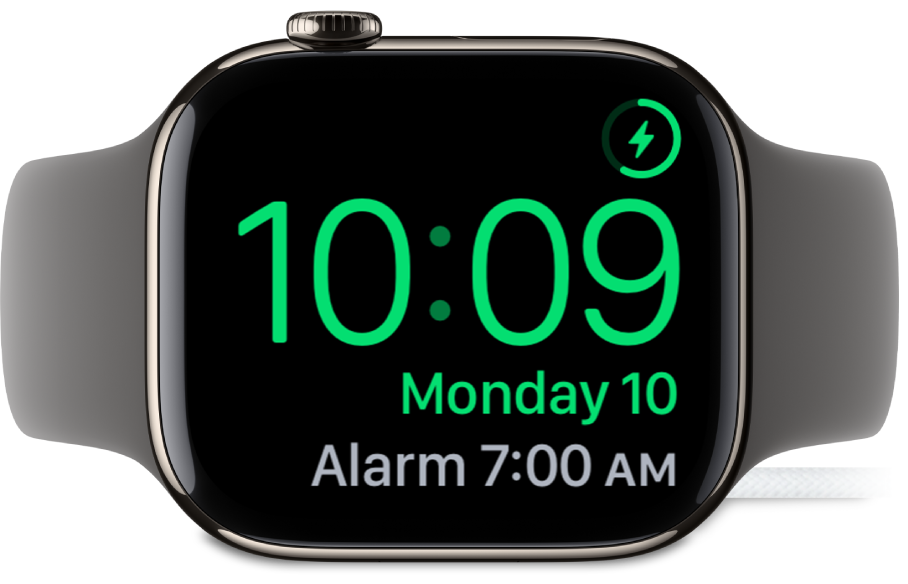 Apple Watch pulkstenis ir novietots uz sāniem un pievienots lādētājam. Tā ekrāna augšējā labajā stūrī ir redzams uzlādes simbols, zem tā ir parādīts pašreizējais laiks, kā arī nākamā modinātāja laiks.