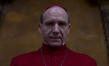 a figure dressed in red clergy attire stands in front of a blurred background