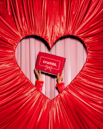 hands holding a crumbl cookie box framed by red fabric in a heart shape