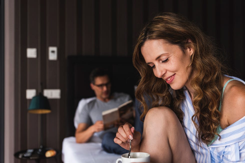 Smiling woman with cup of coffee in bedroom with man in background