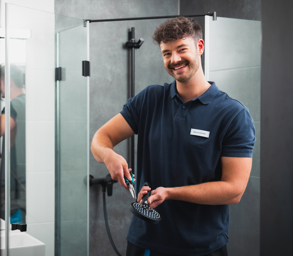 Smiling maintenance technician repairing a showerhead with pliers in a modern bathroom.