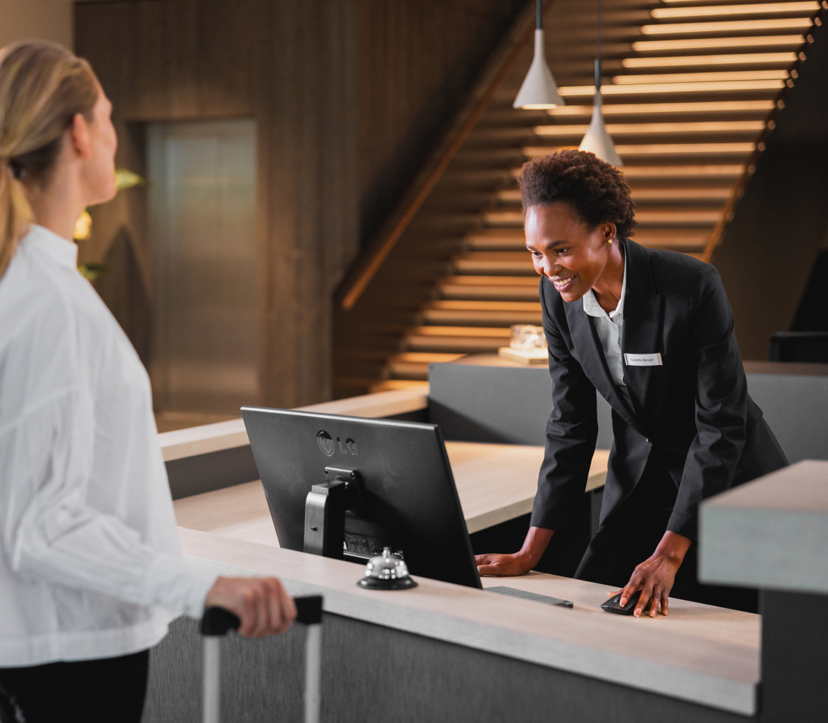 Smiling hotel receptionist in a black blazer assisting a guest at the front desk, with the guest holding the handle of a suitcase. The modern lobby features a staircase and pendant lighting in the background.