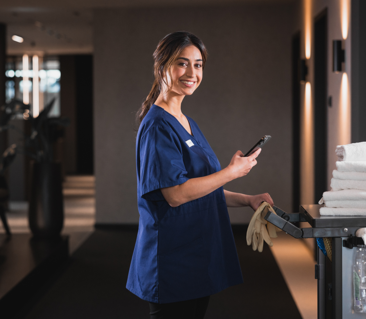 Smiling housekeeping staff member standing beside a cleaning cart, holding a smartphone in a dimly-lit hallway.