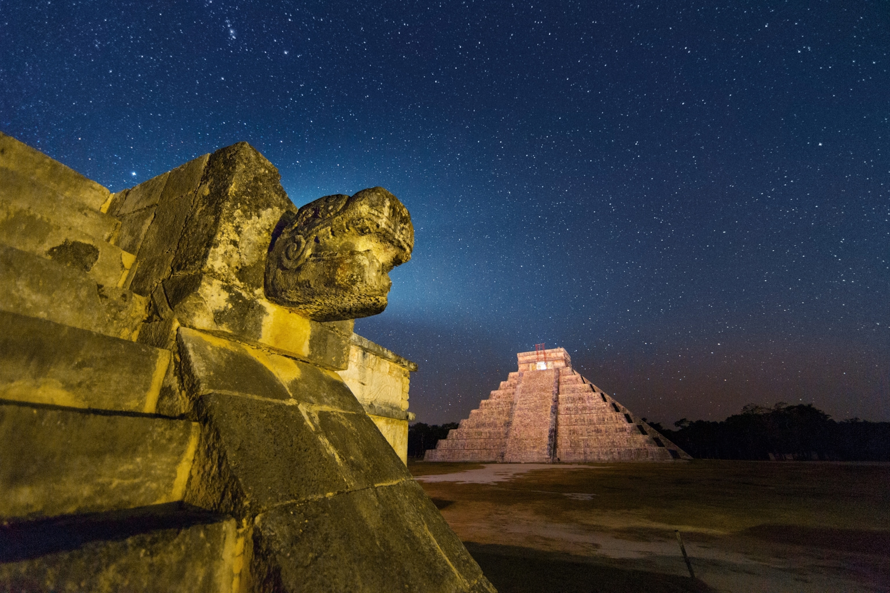 A pyramid and a platform adorned with a feathered serpent's beneath a star filled sky.