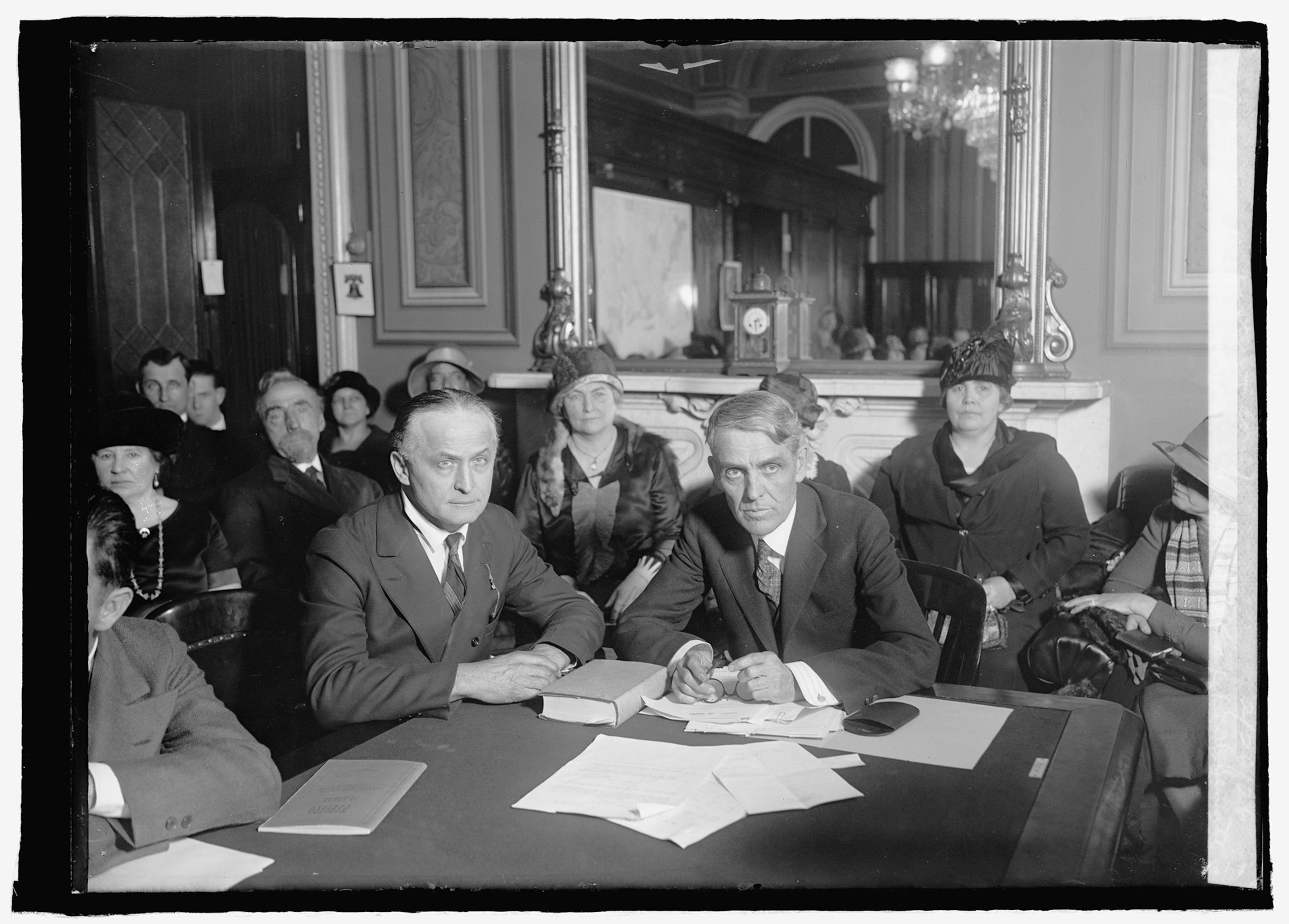 A black and white photo from 1926 with two men and suits sitting at a table and other people in coats and hats sitting around them in luxurious room.