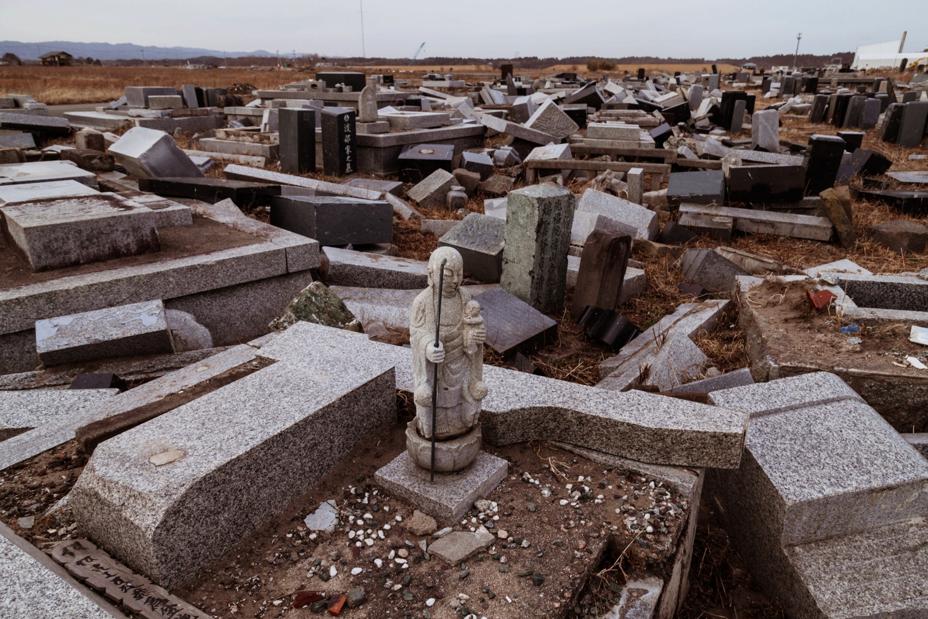 A small statue of Buddha stands amongst damaged gravestones scattered across the ground.