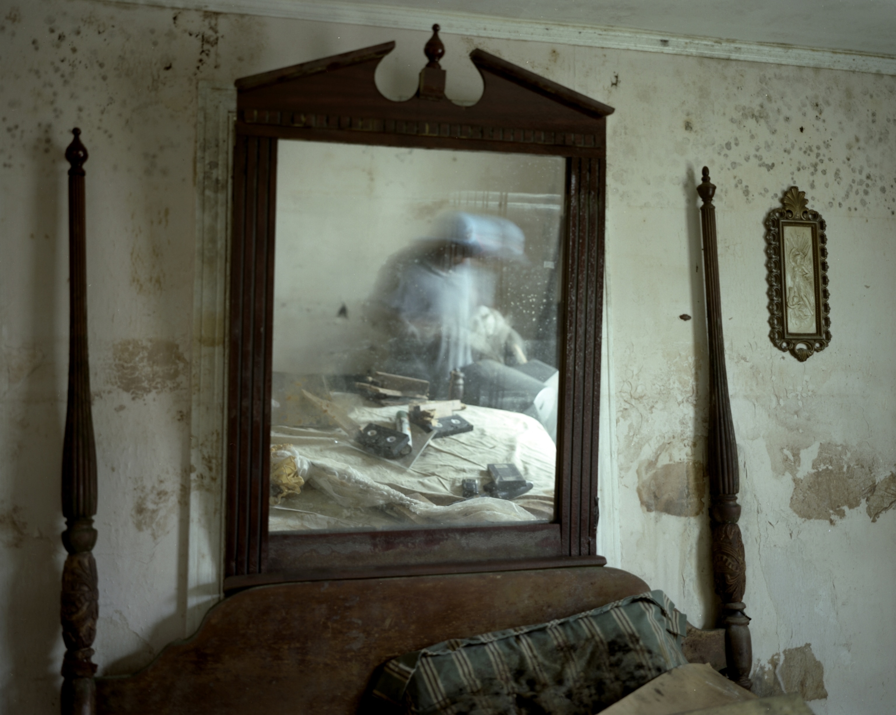 A flood-damaged bedroom. Wooden rails of a headboard frame a wooden mirror, which reflects the blurred image of someone sorting through the rubble.