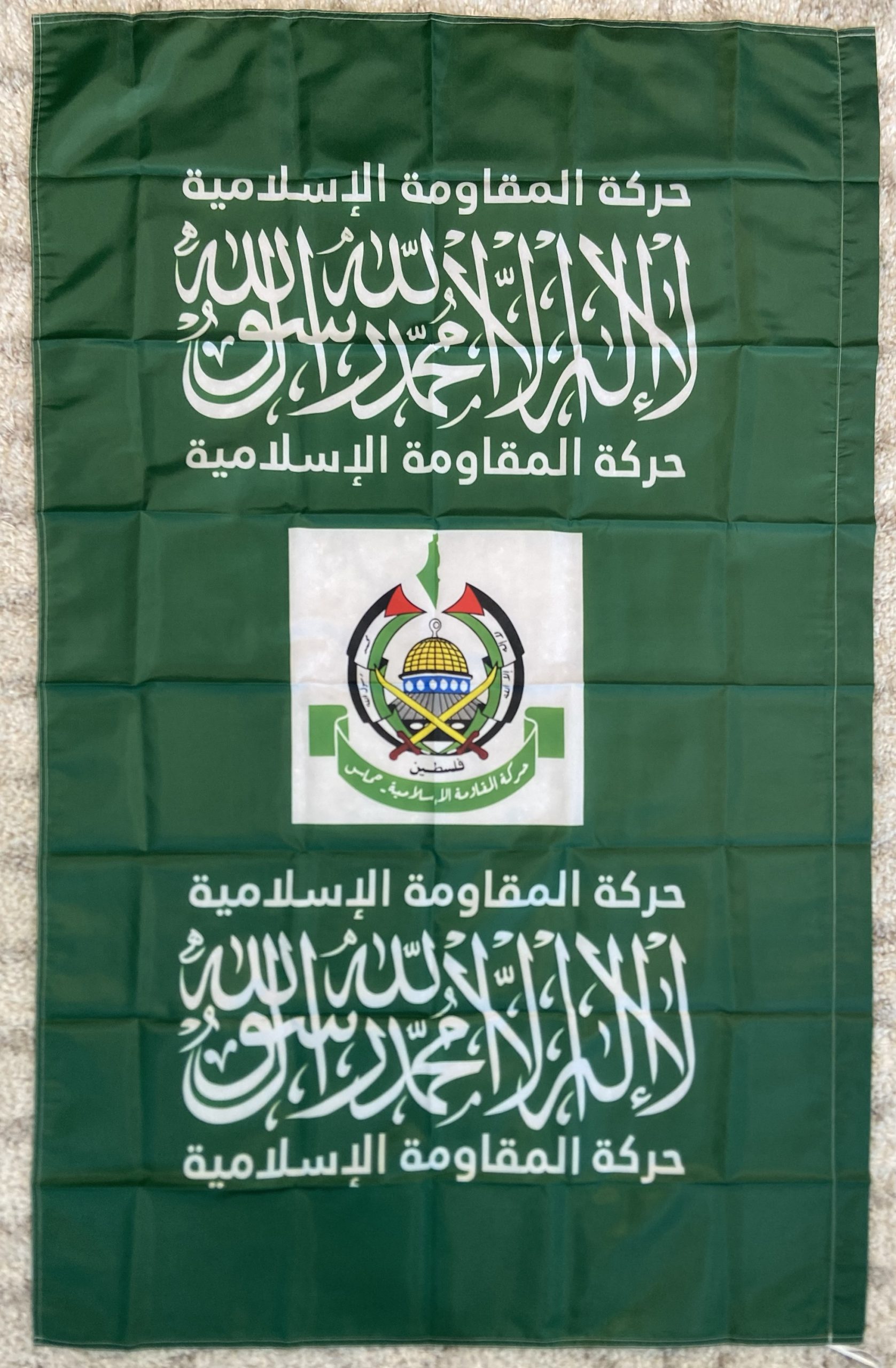 This image showcases the flag of Hamas, a prominent Palestinian political and militant organization. The flag features a dark green background with the Shahada, the Islamic declaration of faith, written in white Arabic script at the center. Below the Shahada is the emblem of Hamas, depicting crossed swords, a globe, and the Dome of the Rock, symbolizing Palestinian nationalism and resistance. Surrounding the central elements are additional Arabic inscriptions. This flag is emblematic of Hamas's identity and its role in the Palestinian-Israeli conflict. This image shows the flag of Hamas, featuring a dark green background with Arabic script in white. At the top center, there is the Shahada, the Islamic declaration of faith. Below the Shahada, there is an emblem featuring crossed swords, a globe, and the Dome of the Rock, symbolizing the Palestinian cause. Additional Arabic text surrounds the central elements, reinforcing messages of faith and resistance. The flag represents the political and militant organization Hamas, which is known for its role in the Palestinian-Israeli conflict.