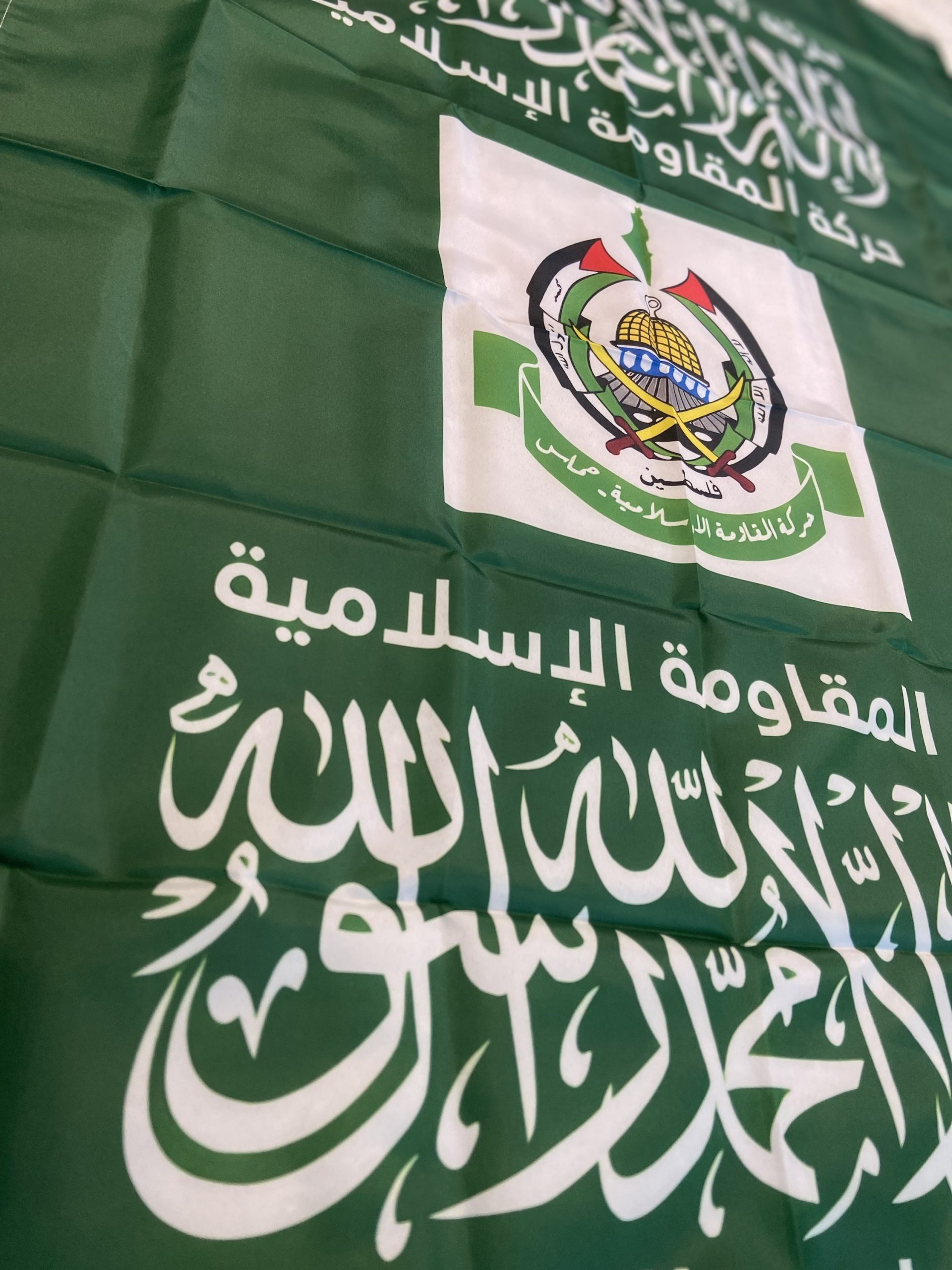 This image showcases the flag of Hamas, a prominent Palestinian political and militant organization. The flag features a dark green background with the Shahada, the Islamic declaration of faith, written in white Arabic script at the center. Below the Shahada is the emblem of Hamas, depicting crossed swords, a globe, and the Dome of the Rock, symbolizing Palestinian nationalism and resistance. Surrounding the central elements are additional Arabic inscriptions. This flag is emblematic of Hamas's identity and its role in the Palestinian-Israeli conflict. This image shows the flag of Hamas, featuring a dark green background with Arabic script in white. At the top center, there is the Shahada, the Islamic declaration of faith. Below the Shahada, there is an emblem featuring crossed swords, a globe, and the Dome of the Rock, symbolizing the Palestinian cause. Additional Arabic text surrounds the central elements, reinforcing messages of faith and resistance. The flag represents the political and militant organization Hamas, which is known for its role in the Palestinian-Israeli conflict.