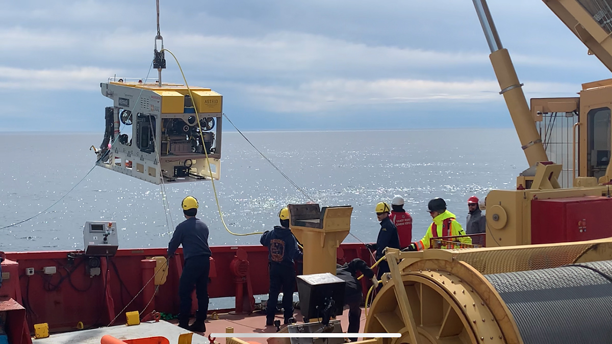 A boxy remotely operated vehicle is lowered by crew in yellow hard hats into the ocean off CCGS Amundsen.