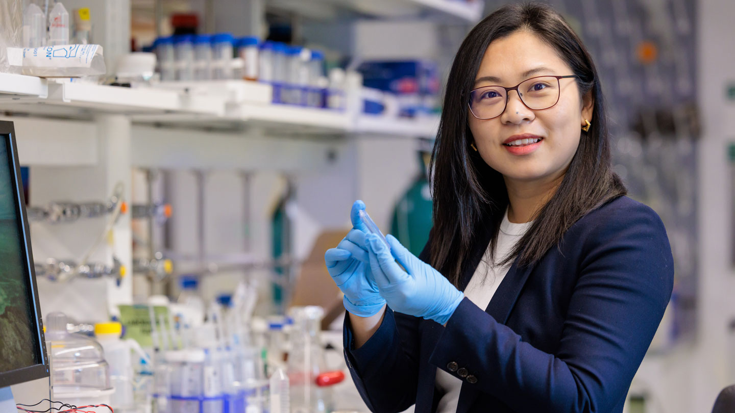 Chong Liu is standing to the right, facing the camera, in a lab. She is wearing a navy jacket and blue nitrile gloves. She has long black hair and wears glasses.